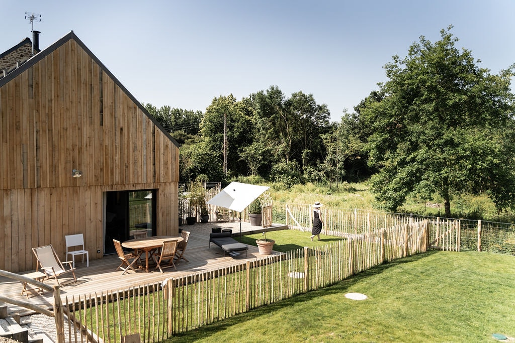 A modern wooden structure is set against a lush green backdrop, with a wooden deck featuring a round dining table and white chairs. A lounge area is visible, along with a sunshade, surrounded by a natural landscape and a fenced yard.