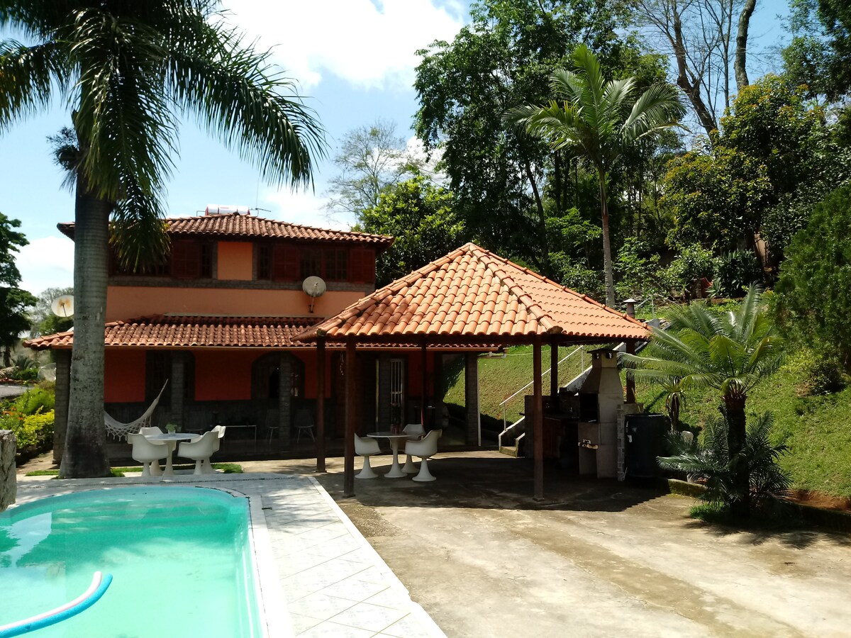 An outdoor area features a private pool with a light turquoise hue, surrounded by palm trees and lush greenery. A pavilion with a terracotta roof houses a grill, positioned next to a spacious patio. The two-story structure is visible in the background.