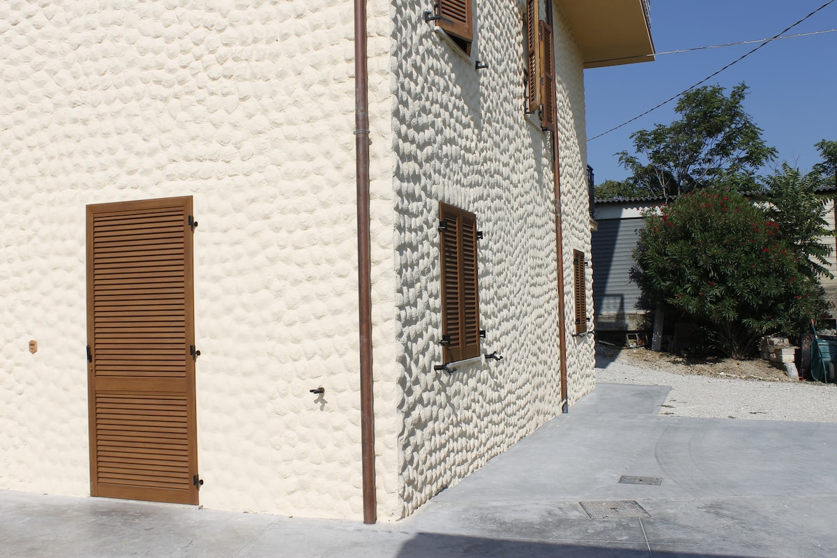 The exterior of a country house is shown, featuring textured light-colored walls and brown wooden shutters. The entrance is highlighted by a door on the left, and the surrounding area includes gravel, greenery, and additional structures in the background.