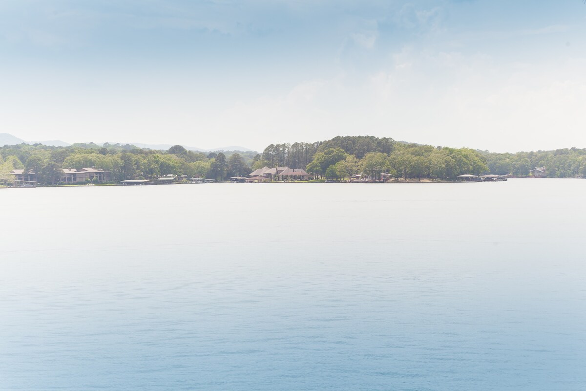 A tranquil lake view features calm waters reflecting soft blue and white hues of the sky. Lush greenery lines the shore, with hints of residential structures nestled among the trees, suggesting a peaceful, serene atmosphere.