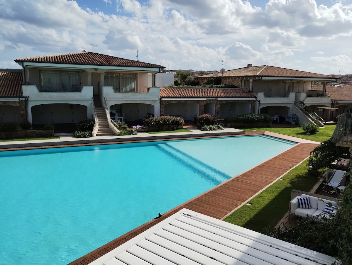 A spacious resort area showcases a large swimming pool with clear blue water and wooden decking. Surrounding greenery and well-maintained gardens feature lounge chairs, and several buildings are positioned nearby, providing easy access to the pool.