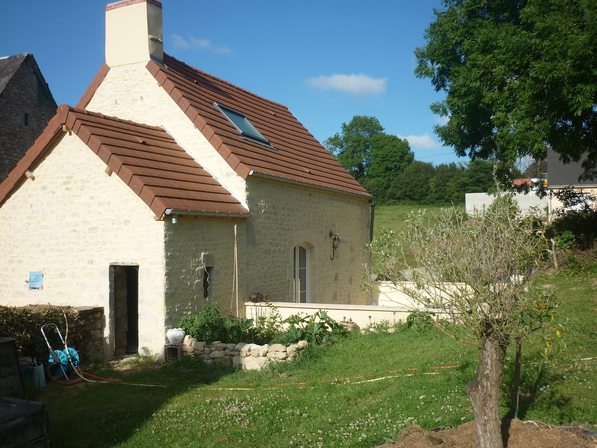 The exterior of a cottage features a stone façade and a sloped roof. A small skylight is visible, and greenery surrounds the structure, providing a serene backdrop. The property is set in a tranquil environment with trees and a neatly maintained garden area.