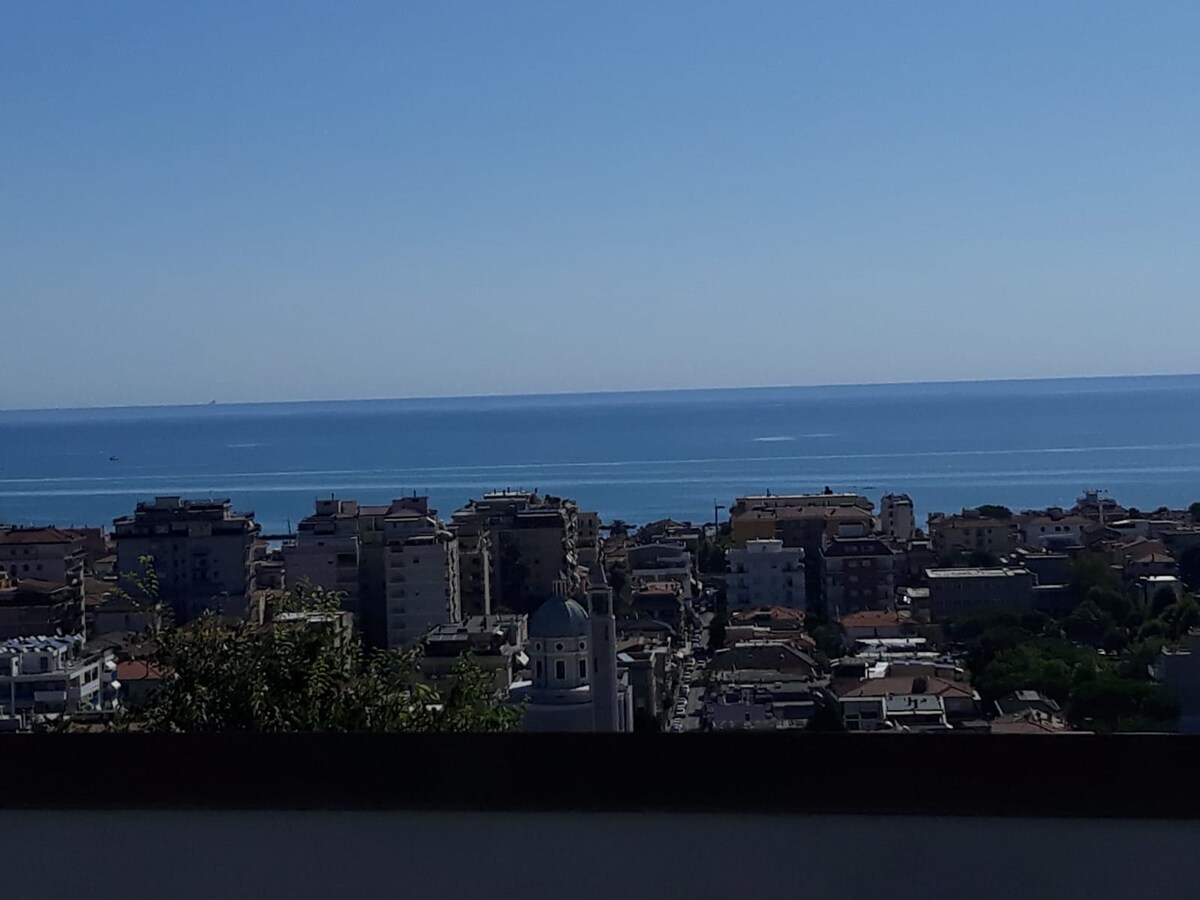 A panoramic view of the coastline is presented, showcasing the sea blending into the horizon. Residential buildings are visible in the foreground, with a mix of architecture and greenery. The sky is clear and blue, enhancing the serene atmosphere of the scene.