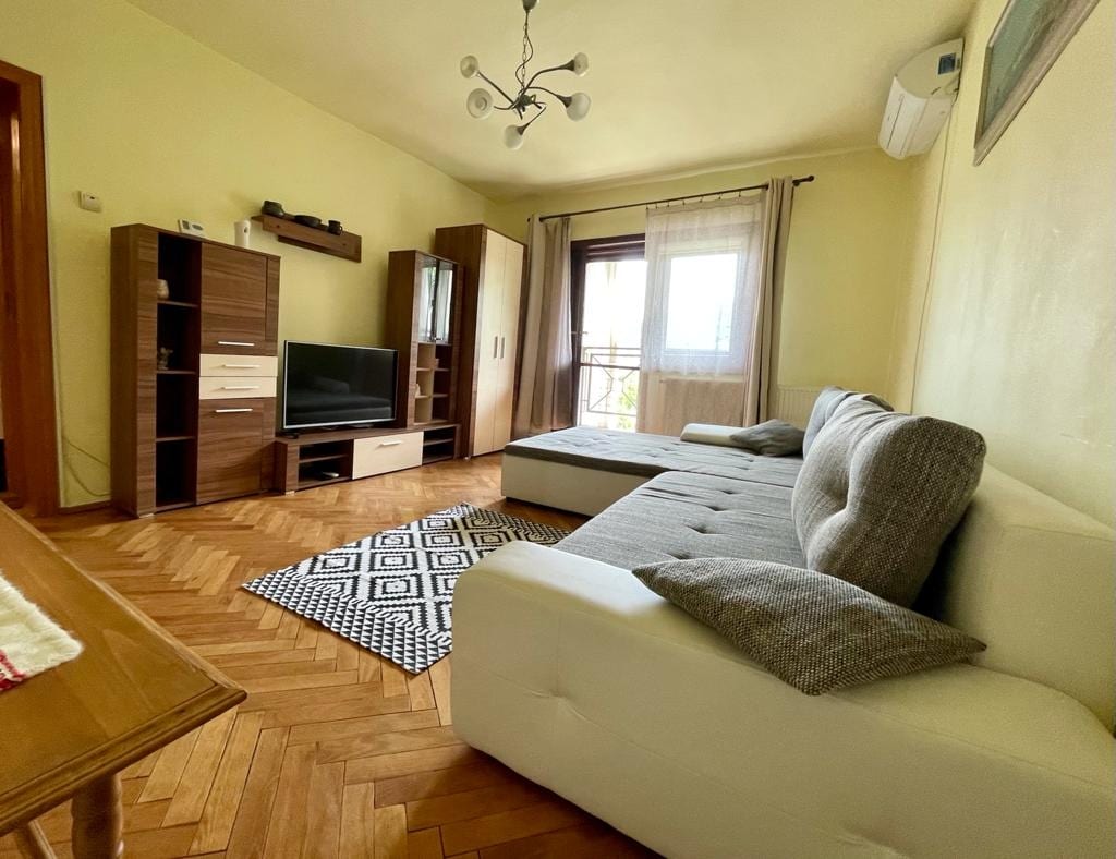 A living area is shown with a light-colored sectional sofa and a patterned rug on the wooden floor. A flat-screen television is positioned on a modern cabinet, while light streams through a window with sheer curtains. An air conditioning unit is mounted on the wall.