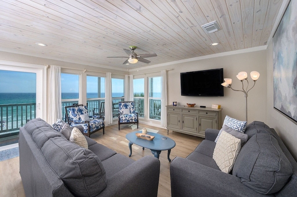 The living area features large windows that provide expansive views of the Gulf. Two plush sofas are arranged around a coffee table, complemented by stylish armchairs. A flat-screen TV is mounted on a light-colored wall, and light-colored curtains enhance the airy atmosphere.
