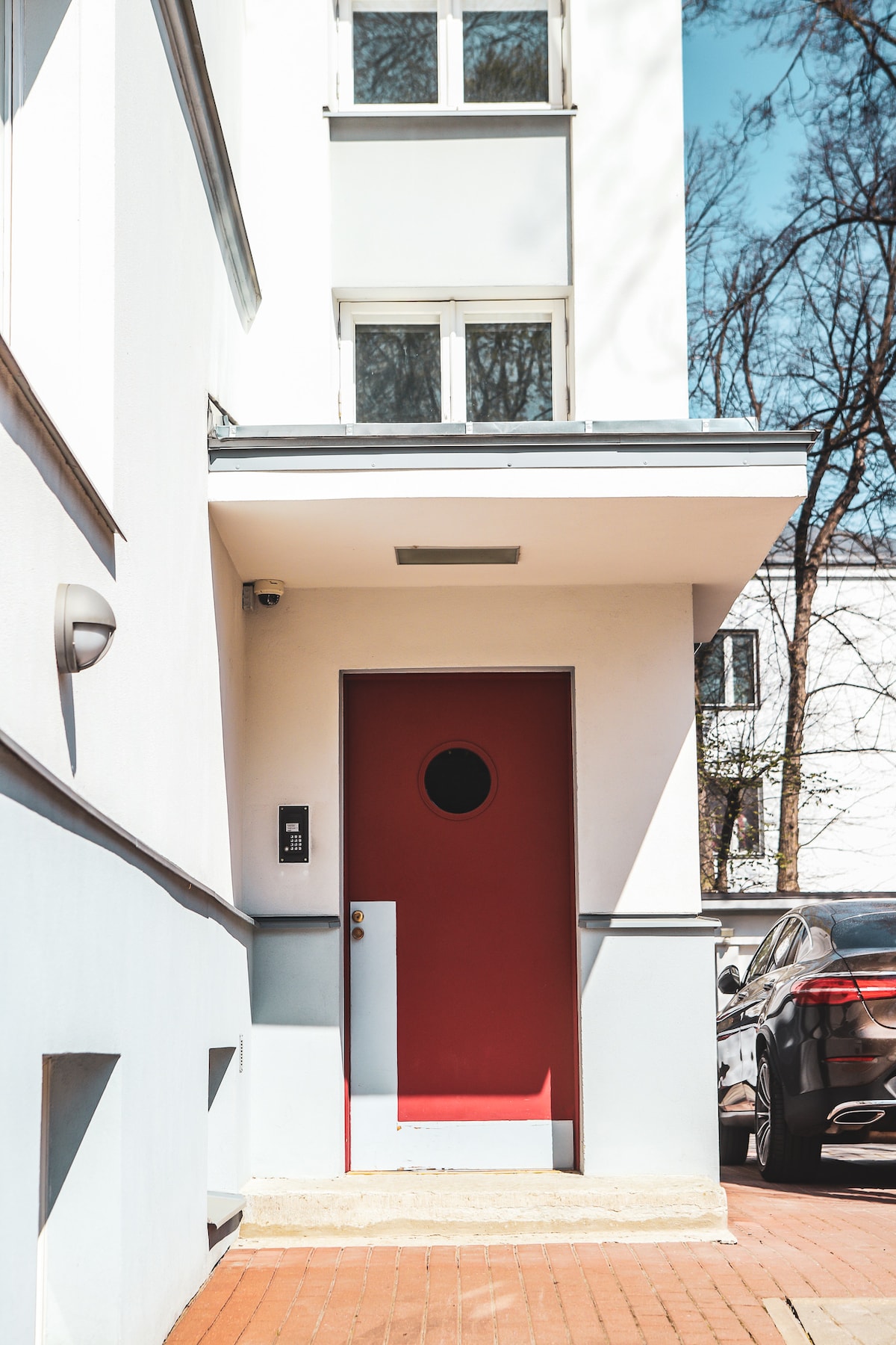 A contemporary entrance features a bold red circular door, framed by a clean white facade. Above, light is provided by a modern fixture. The surrounding area is paved, with a vehicle parked nearby, contributing to the urban setting.