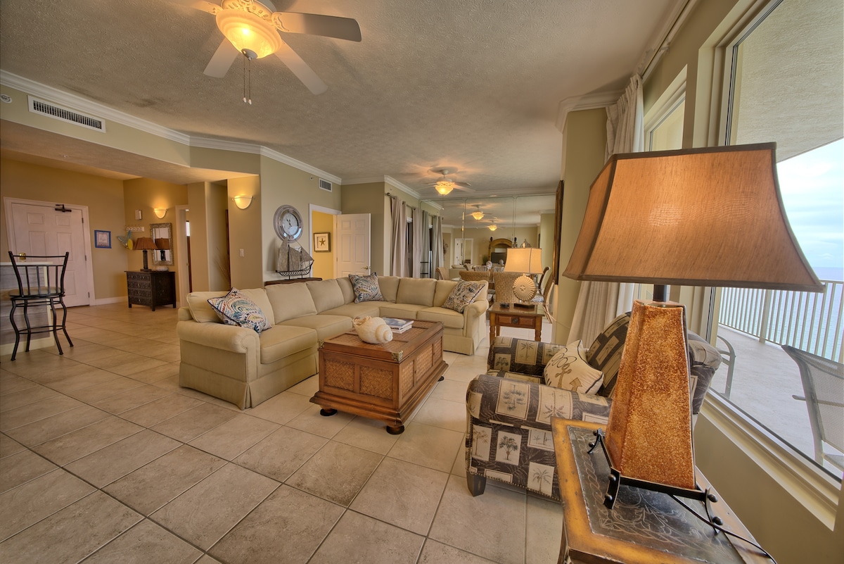 The spacious living area features large windows that provide ample natural light and captivating views of the ocean. A light-colored sectional sofa is positioned in the center, complemented by a decorative coffee table and various accent chairs. A lamp adds functional lighting to the inviting space.