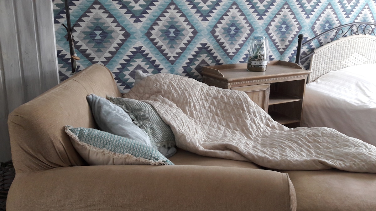 A spacious couch is adorned with soft pillows and a light quilt, inviting relaxation. A bedside table stands nearby, featuring a decorative glass candle holder. The backdrop showcases a patterned wall in shades of blue, enhancing the cozy ambiance of the space.