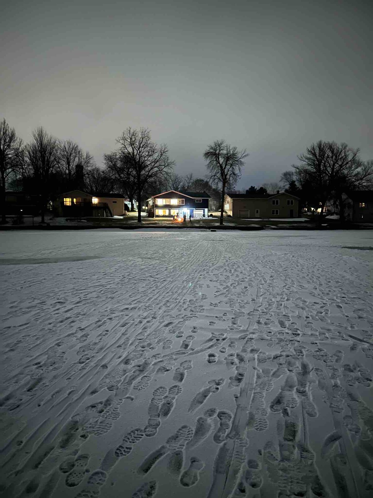 A lakeside home is illuminated against a nighttime backdrop, surrounded by a snowy landscape. Footprints in the snow lead toward the house, which features large windows. Nearby trees are silhouetted against the evening sky, adding to the serene winter atmosphere.