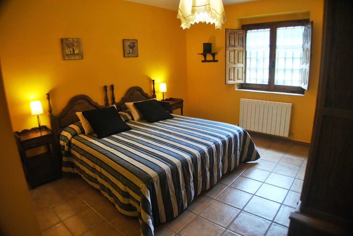 A private double room features a spacious bed covered with a striped blanket. Two bedside lamps provide soft illumination. The warm yellow walls complement the wooden accents, while a window with wooden shutters allows natural light to fill the room, enhancing the overall ambiance.