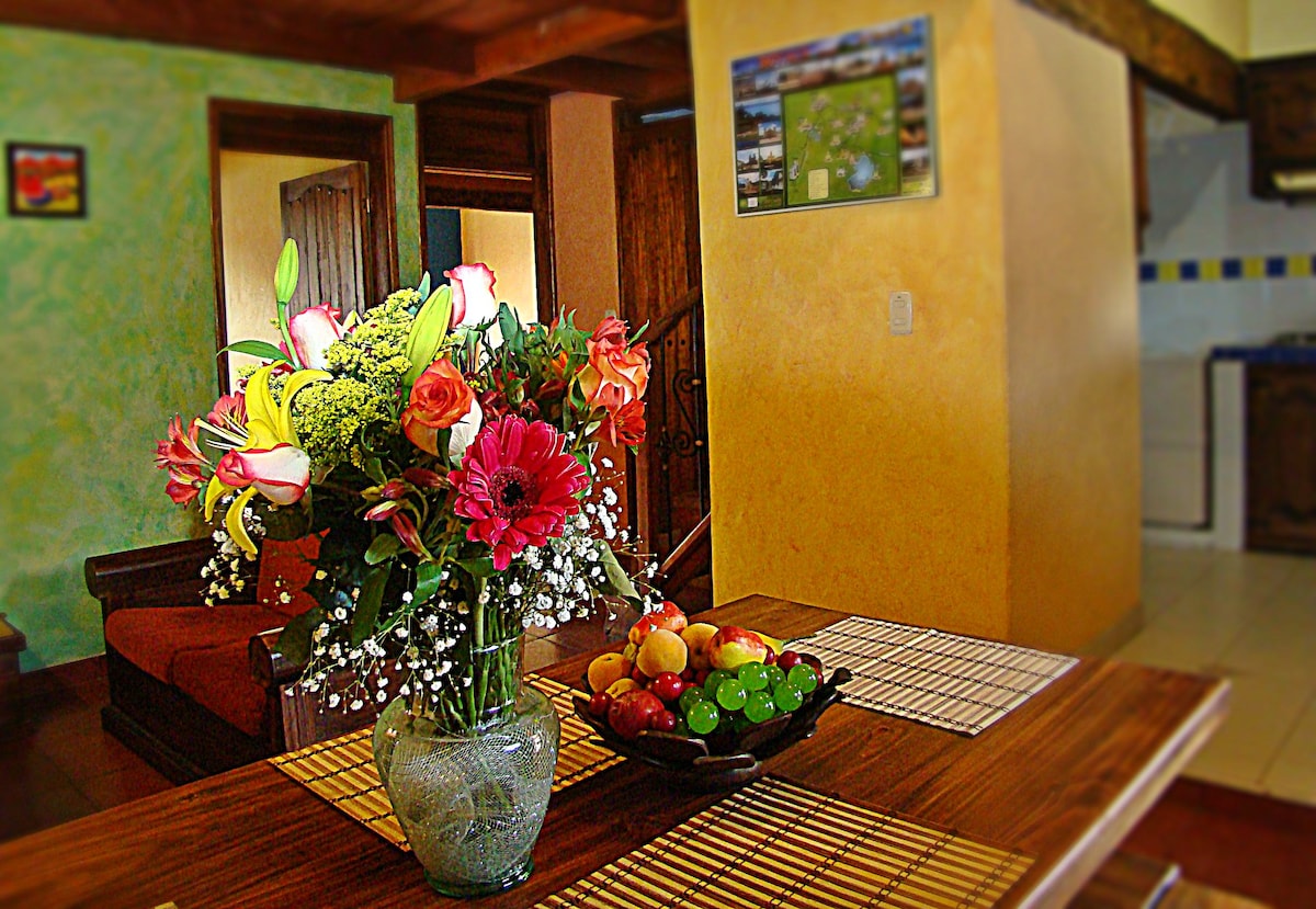 A dining table is arranged with a decorative fruit bowl and a vibrant flower arrangement. The background features a cozy seating area and a glimpse of a kitchen space, all under warm lighting that enhances the rustic furnishings.