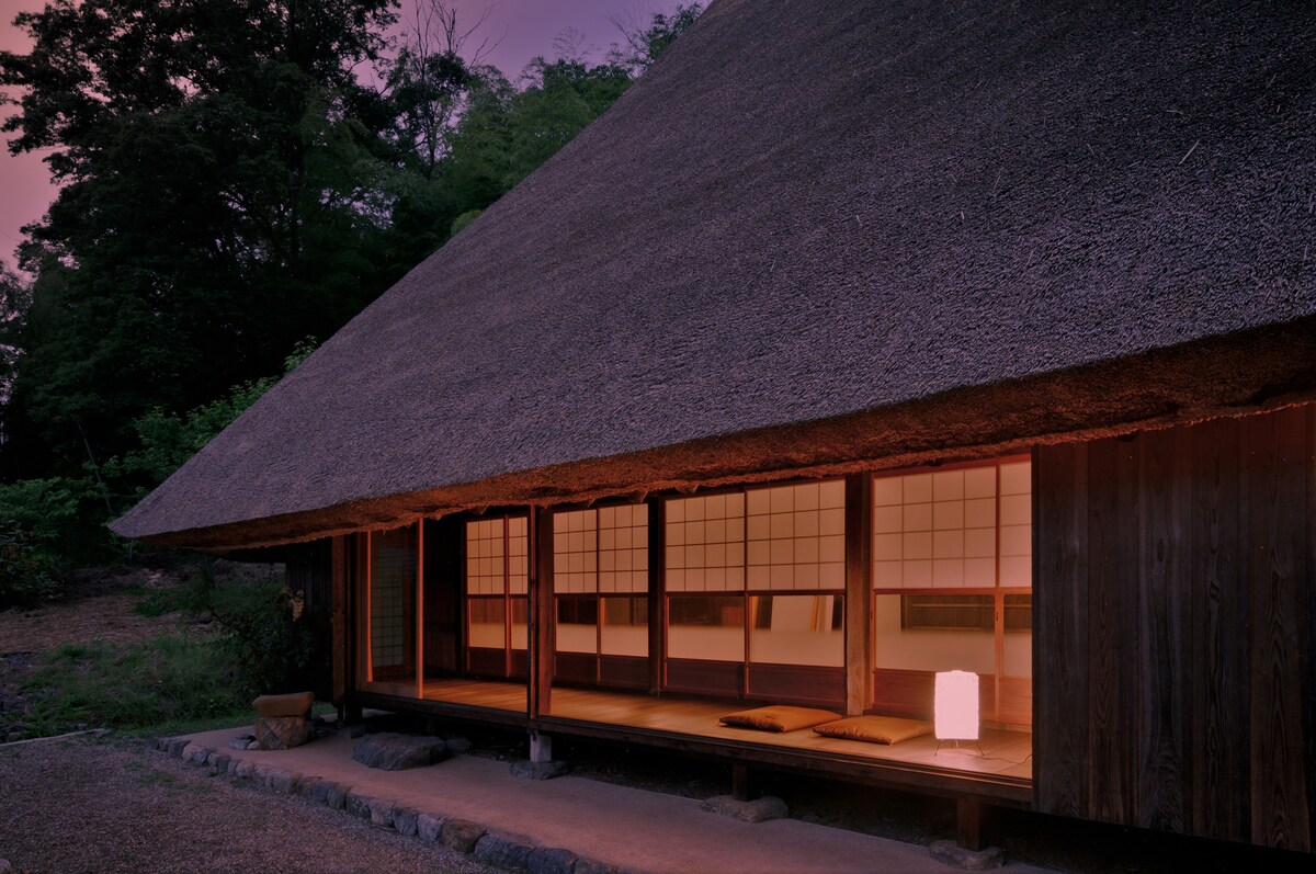 A traditional thatched cottage is shown with a sloping roof and wooden façade. The entrance features sliding doors that open to a wooden porch, accented by tatami mat seating. Soft lighting from within and surrounding greenery create a serene outdoor setting during dusk.