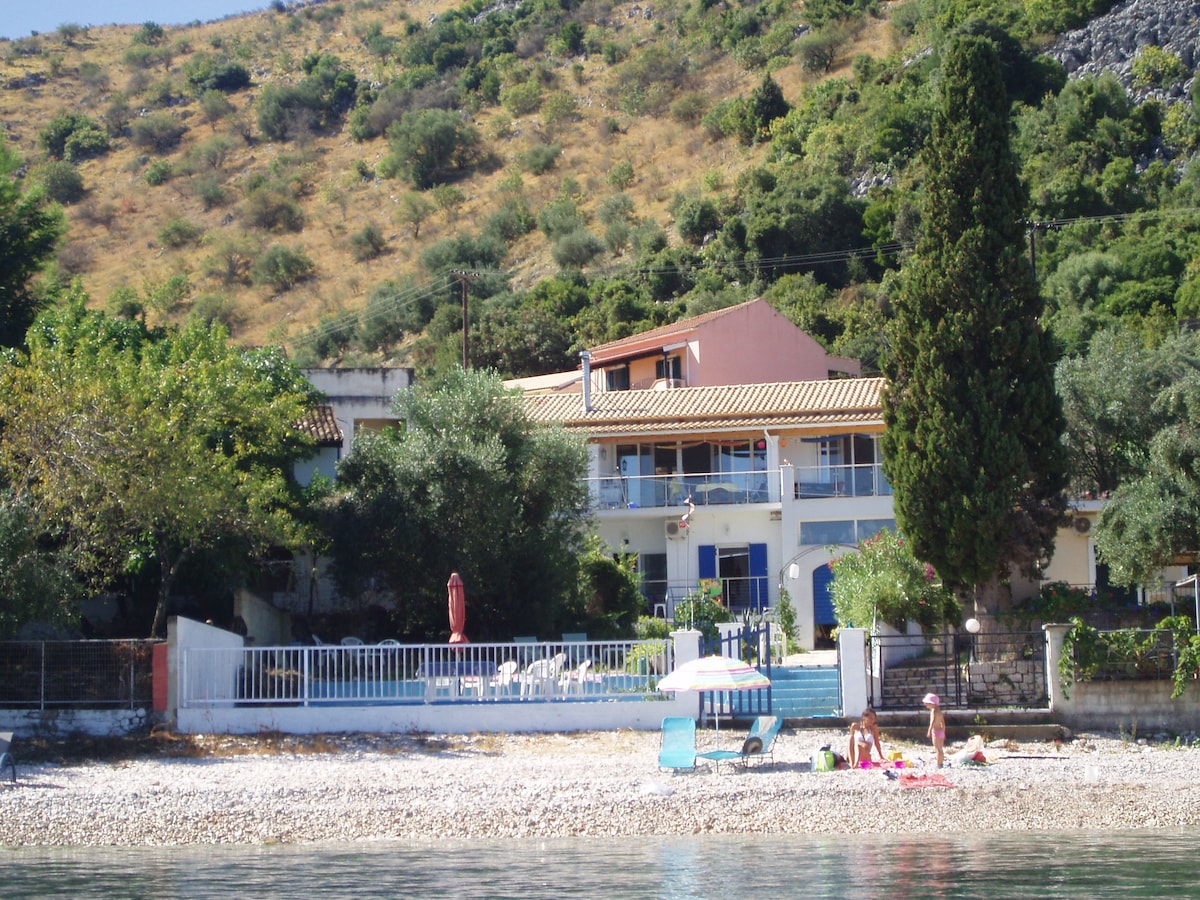 The beach house is set against a backdrop of gently sloping hills, with a garden containing olive trees and beach loungers visible in the foreground. A spacious balcony is seen on the upper level, offering views of the beach and surroundings.