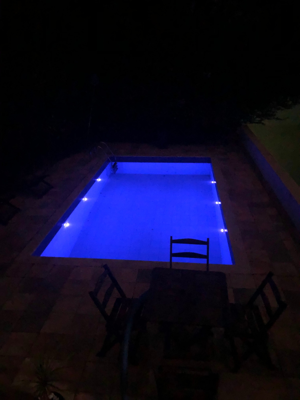 A serene swimming pool is viewed from above, illuminated in soft blue lights. Surrounding the pool are simple wooden chairs and a small table, set against a dark backdrop of foliage. The tranquil water reflects the warm tones of the surrounding outdoor area.