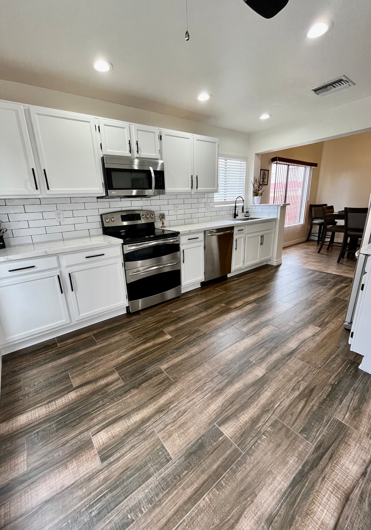 A well-equipped kitchen features modern stainless steel appliances, including a stove, microwave, and dishwasher. White cabinetry contrasts with the dark wooden flooring. Natural light enters through a window, while a dining area is visible through an open doorway.