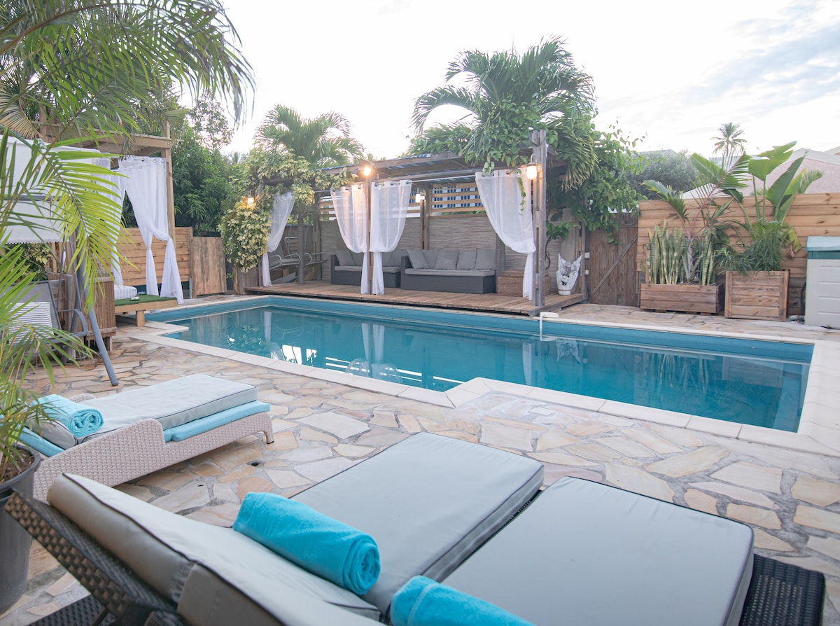 A private outdoor space features a serene pool framed by natural stone tiles, with lounge chairs and towels neatly arranged nearby. A shaded seating area, adorned with curtains and comfortable cushions, adds a cozy touch. Lush greenery enhances the tranquil atmosphere.