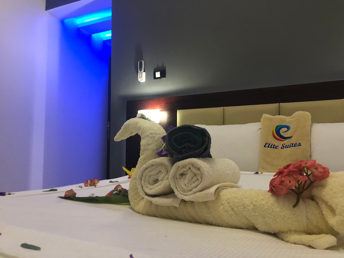 A neatly arranged bed features plush pillows and soft linens, with a decorative towel swan at the forefront. Roll towels are stacked nearby, accompanied by vibrant artificial flowers, adding a touch of color. Subtle blue lighting enhances the room's ambiance.