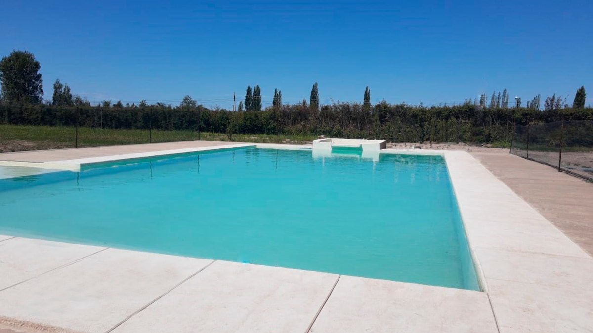 A serene outdoor swimming pool is surrounded by a large, paved area. The crisp blue water reflects the clear sky, with lush greenery visible in the background. The pool features a gentle entry slope, enhancing accessibility.
