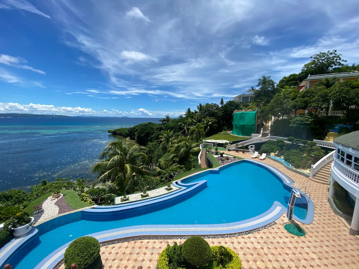 An expansive infinity pool offers uninterrupted views of the sea and lush greenery. Palm trees are positioned around the pool area, with sun loungers placed along the water's edge. The sky reflects various shades of blue, enhancing the serene environment.