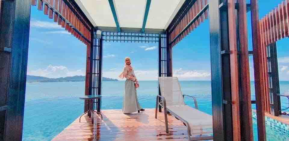 A spacious wooden deck extends over the water, featuring a pair of loungers with cushions. A shaded area provides comfort, while the ocean and distant hills are visible in the background under a clear sky.