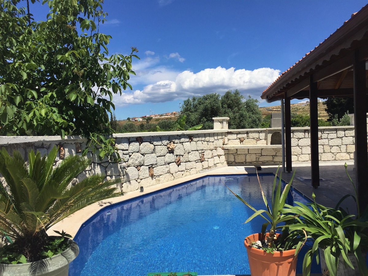 A serene outdoor space features a clear blue swimming pool surrounded by lush greenery. The area is framed by a natural stone wall and a shaded structure, providing an inviting retreat. Scenic views of the landscape extend beyond the pool, enhancing the tranquil atmosphere.
