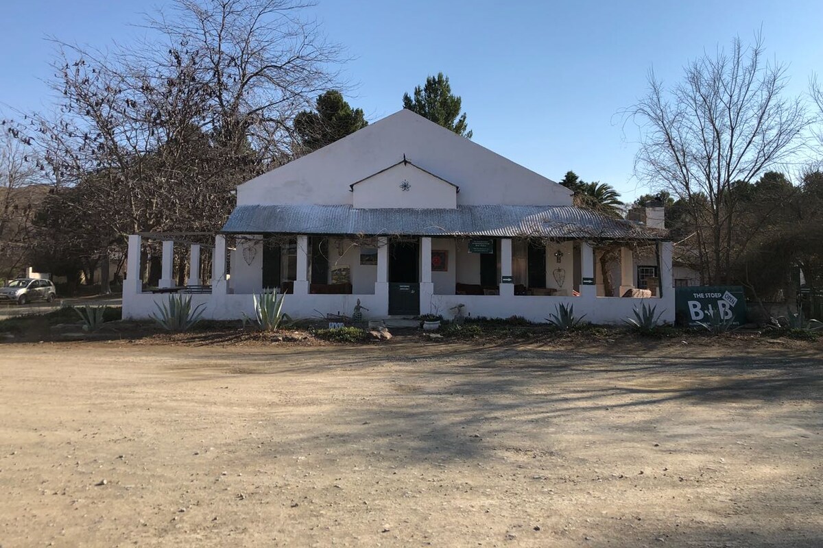 A traditional white-walled building with a gabled roof is presented, surrounded by gravel pathways and sparse vegetation. Large windows line the facade, and a welcoming sign indicating 'The Stoep B&B' is visible. Leafless trees create a rustic backdrop against a clear blue sky.