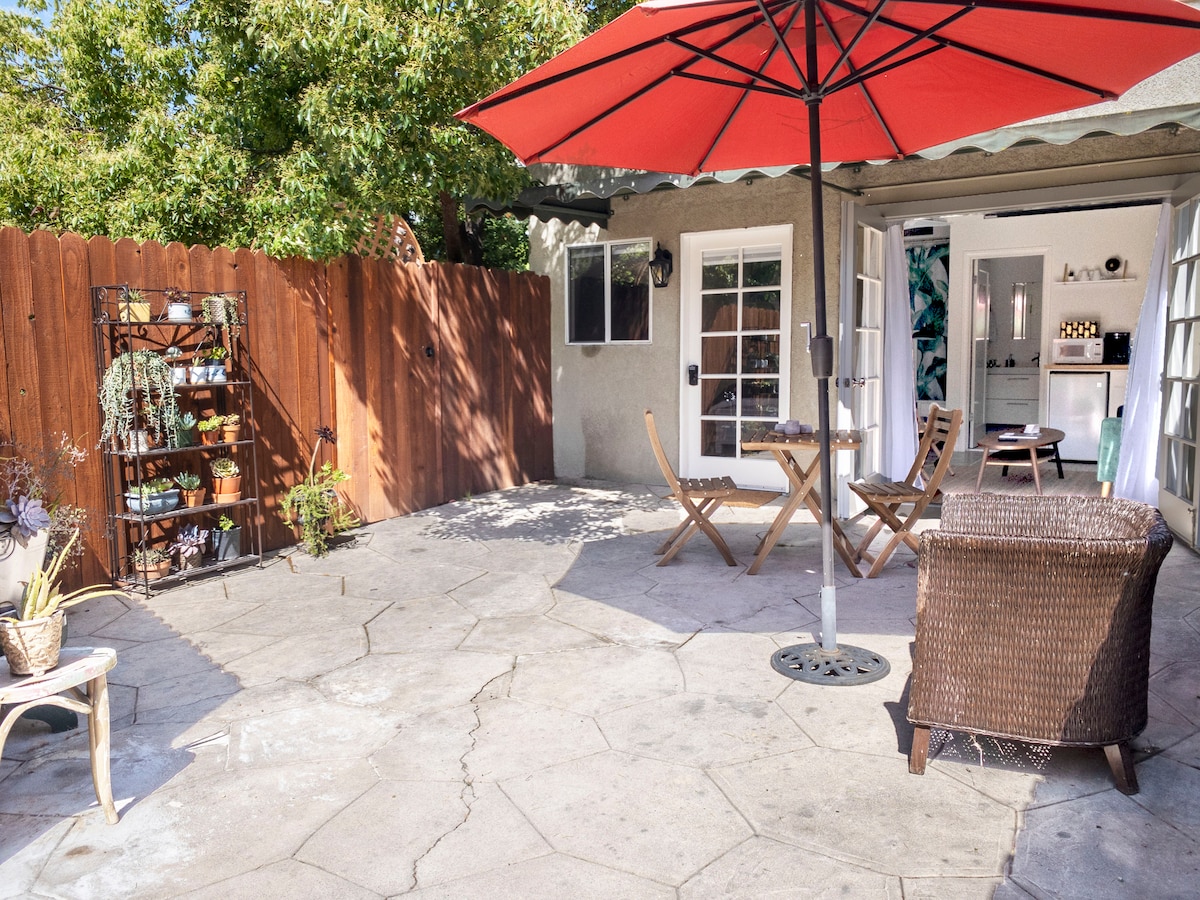 A private patio area features a round table surrounded by two folding chairs under a large red umbrella. A wicker lounge chair is positioned nearby on the textured concrete surface. A wooden privacy fence is visible, along with a small plant rack holding potted plants.