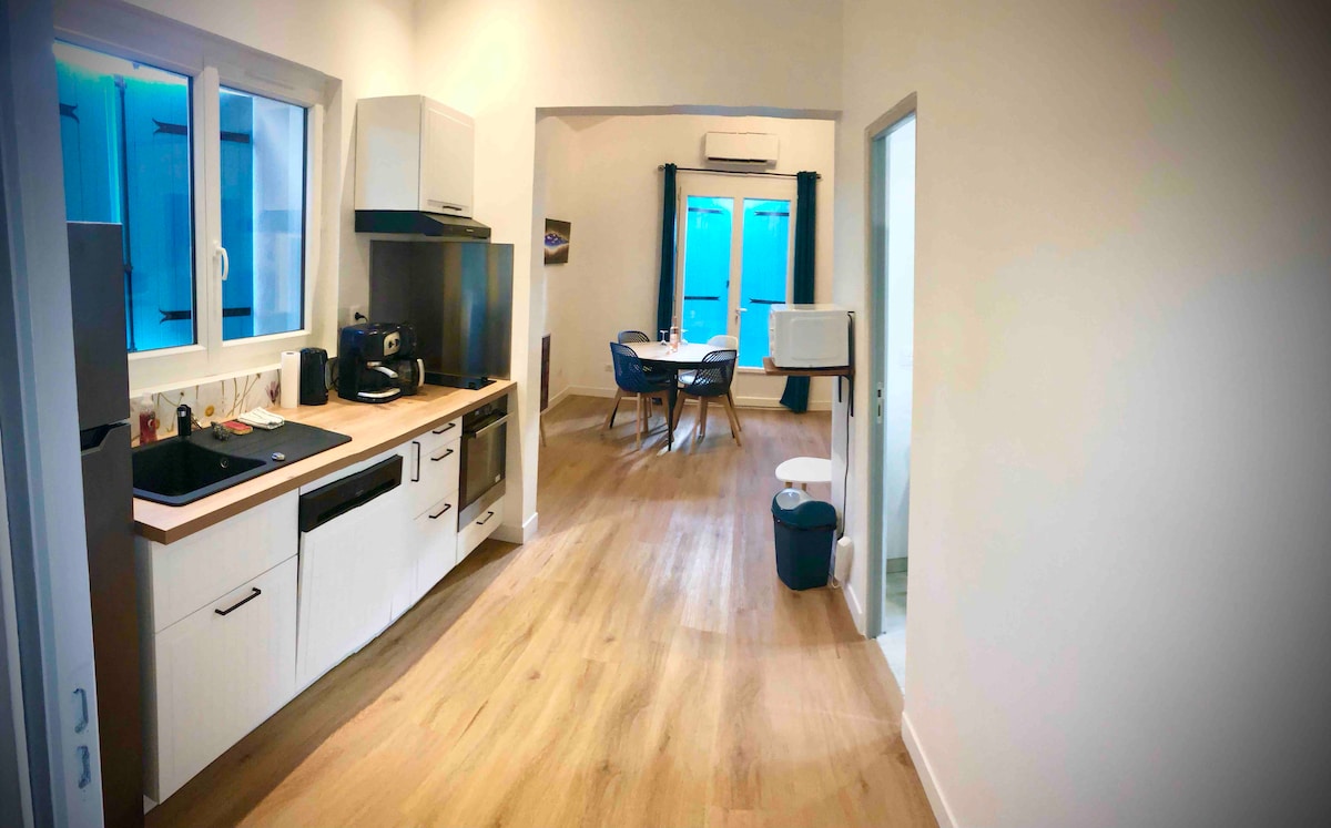 A modern kitchen area is equipped with a range of appliances, including a stove and a microwave. The space features light wood flooring and a dining table with chairs, illuminated by natural light from large windows that provide a view of the outdoors.