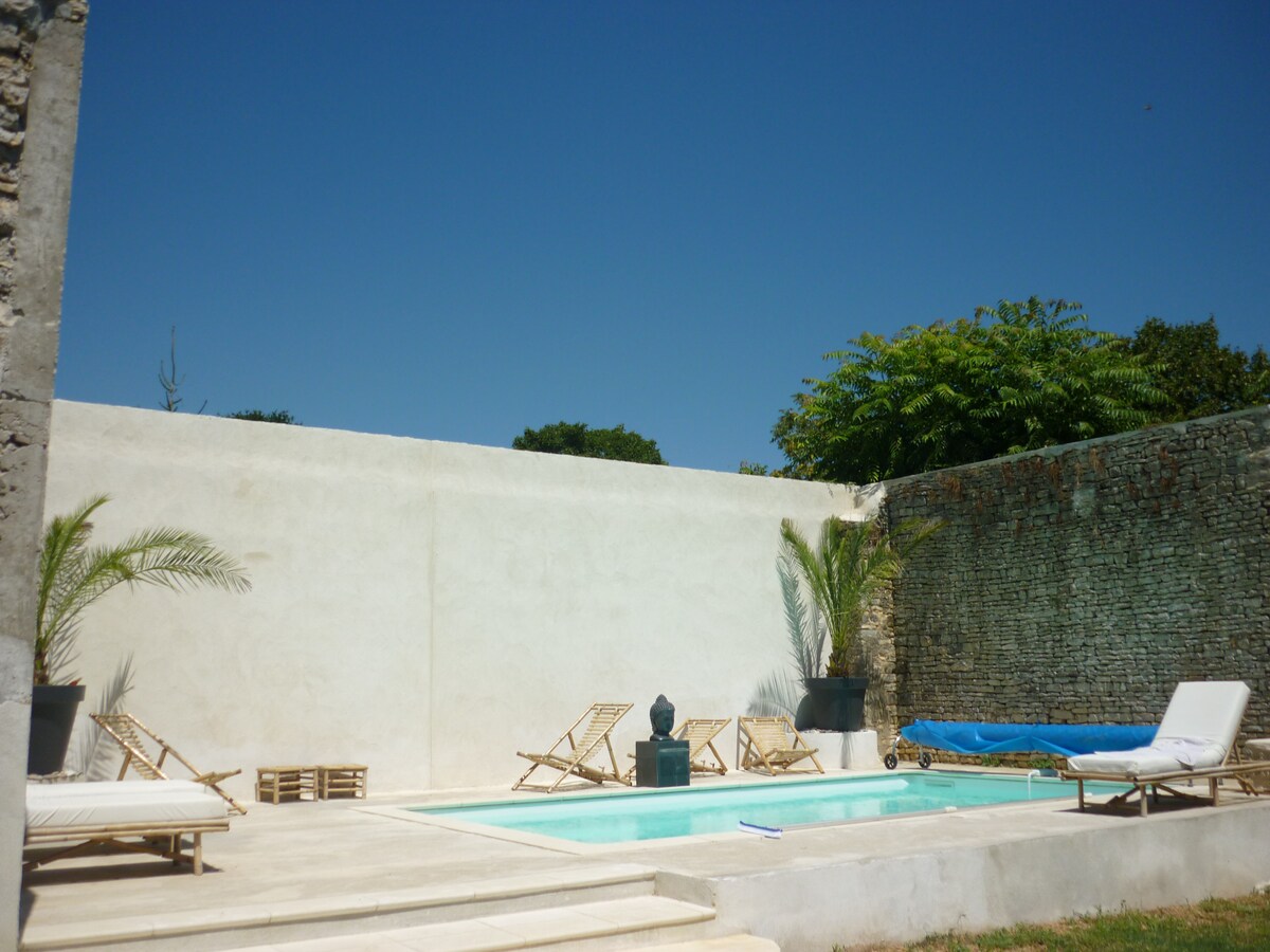 A private pool area is featured, surrounded by a stone wall and lush greenery. Two loungers are positioned beside the pool, while a potted palm adds a touch of nature. Sunlight brightens the space, creating a peaceful atmosphere for relaxation.