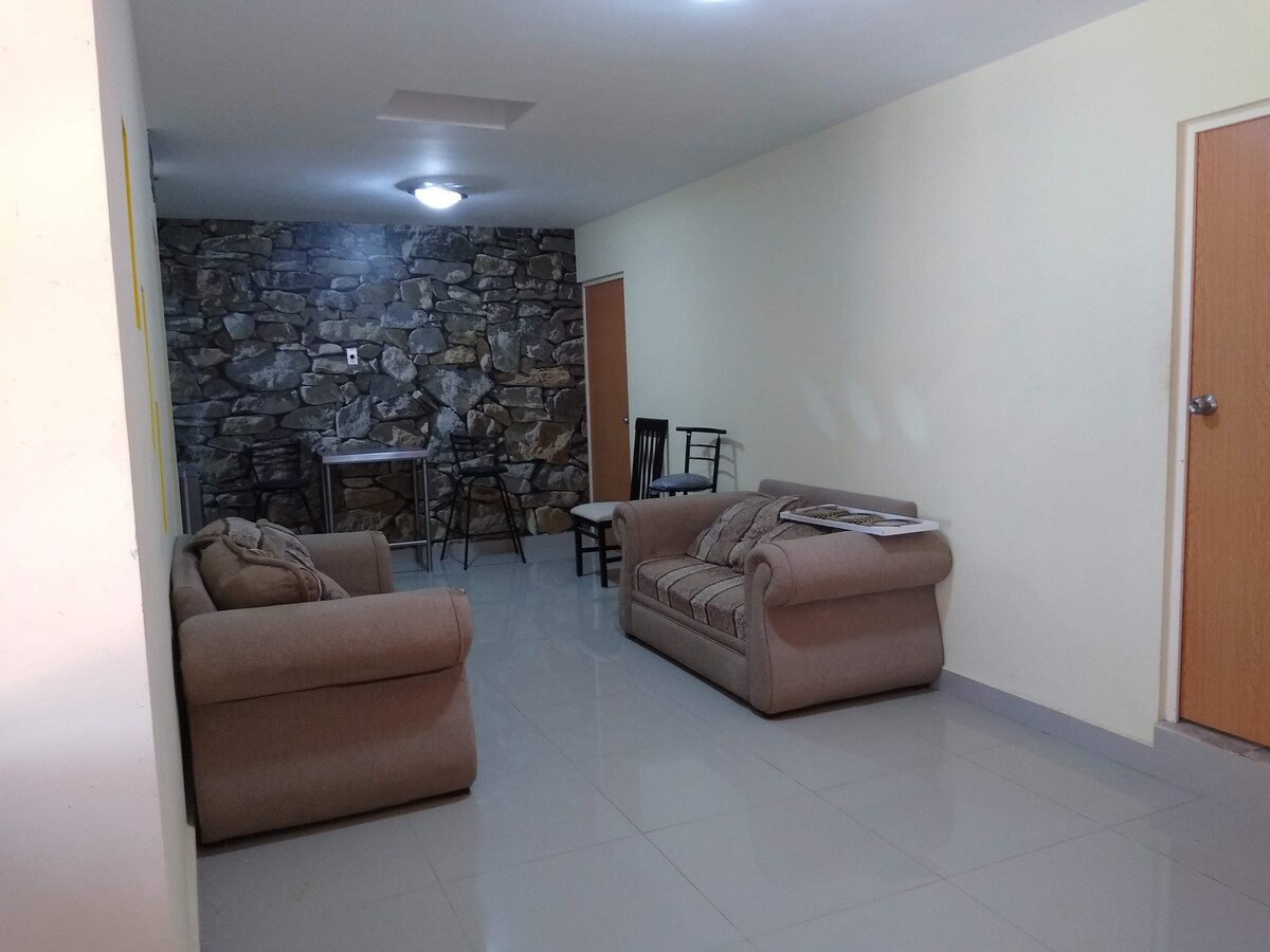 The living area features a stone accent wall, complemented by two comfortable sofas and a small dining table surrounded by chairs. Natural light is present, illuminating the tiled floor and creating a bright and open space.