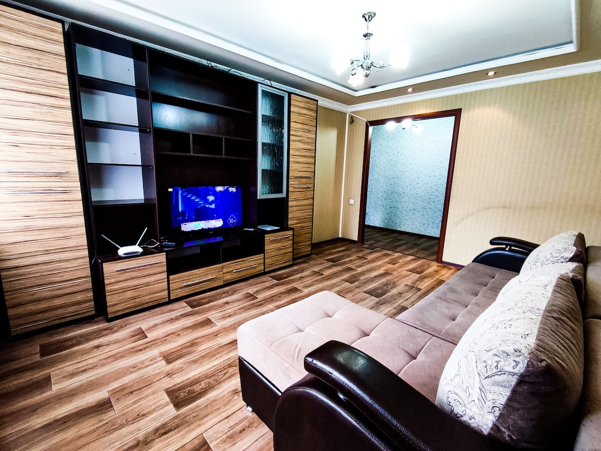 A living area features a spacious sectional sofa complemented by decorative cushions. A television is mounted on a modern wooden unit beneath a subtle chandelier. The floor showcases light wooden planks, while walls display a warm beige tone, enhancing the room's inviting layout.