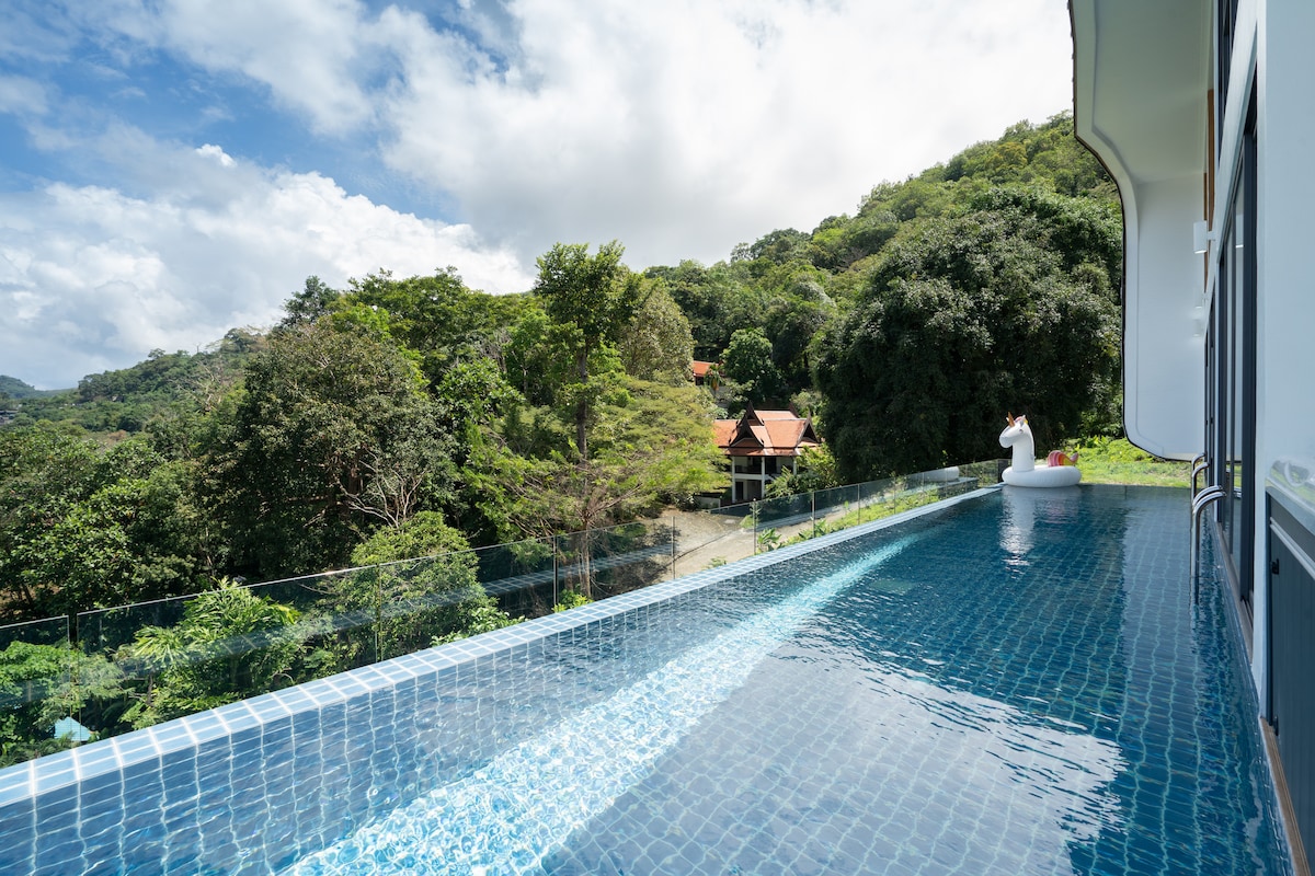 An expansive infinity pool extends along the villa's edge, offering stunning views of the lush landscape and distant structures. The water reflects the clear blue sky, while a gentle breeze rustles the surrounding greenery. A serene atmosphere is created by the private setting.