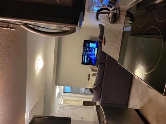 A contemporary kitchen is visible, featuring a stovetop and modern appliances. Beyond, a living area is seen with a gray sofa and a flat-screen television against a light-colored wall. Doorways lead to additional rooms, enhancing the spacious layout.