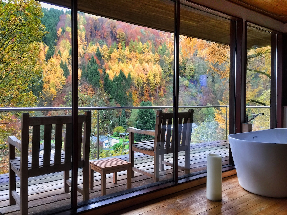 A view of a private balcony features comfortable wooden seating that overlooks a vibrant landscape of autumn foliage. Natural light illuminates the space, and a freestanding bathtub is positioned nearby, enhancing the connection to the surrounding nature.