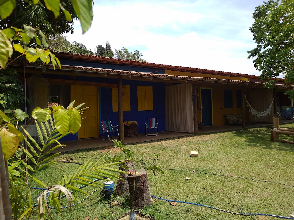 A vibrant exterior of the bungalow is presented, featuring a blue facade adorned with yellow shutters. Relaxing chairs are positioned on a shaded patio, surrounded by greenery. The roof provides shelter, and a hammock can be seen, enhancing the connection to the natural surroundings.