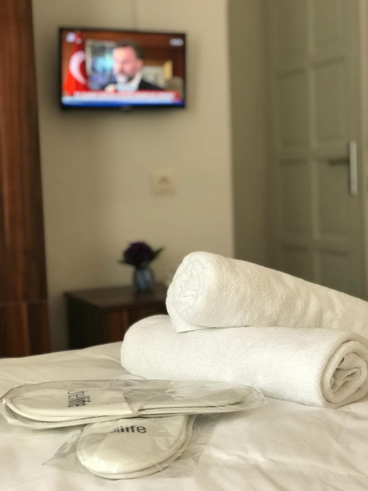A comfortable bedroom setting features neatly arranged white towels and complimentary slippers on a neatly made bed. A small vase with a flower is visible in the background, along with a television showing a news broadcast.