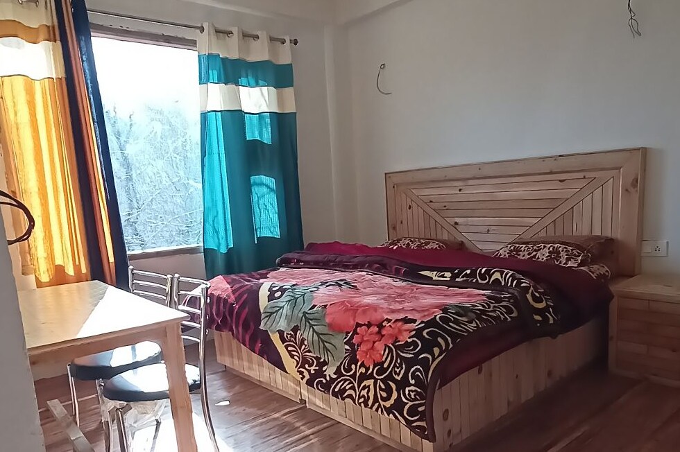 A well-lit bedroom features a spacious bed adorned with a floral blanket. Two large windows are dressed in vibrant curtains, allowing natural light to fill the room. A wooden table and chairs are positioned nearby, enhancing the functional layout.