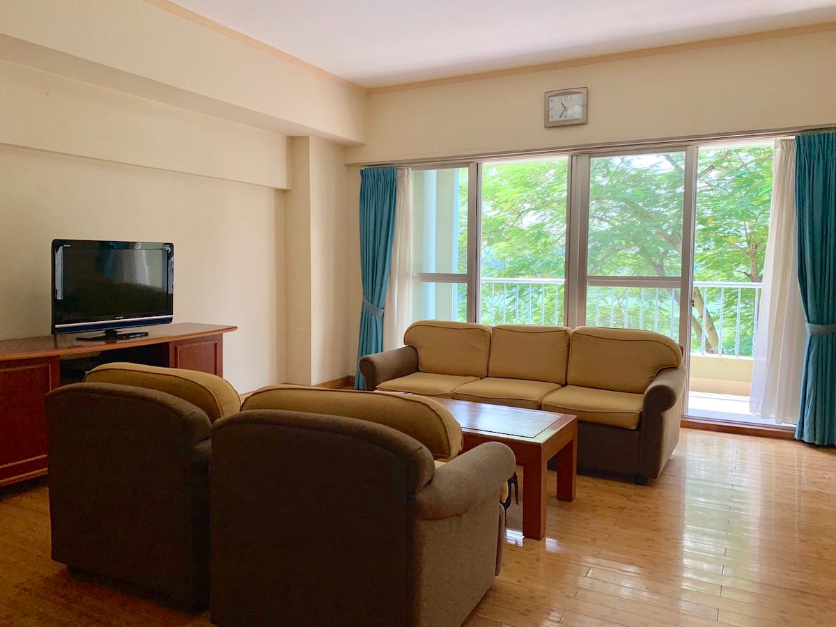 A spacious living area is furnished with a comfortable sofa set and a wooden coffee table. Natural light enters through large windows, flanked by soft blue curtains. A television is positioned on a wooden cabinet against the wall. Greenery can be seen outside the windows.