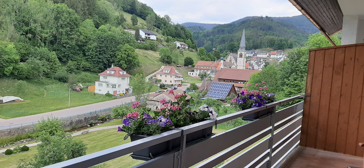 A balcony view showcases a scenic valley filled with lush greenery and residential buildings. Colorful flower boxes line the railing, adding a touch of beauty, while the distant village features a charming church steeple and rooftops partially shaded by the surrounding hills.