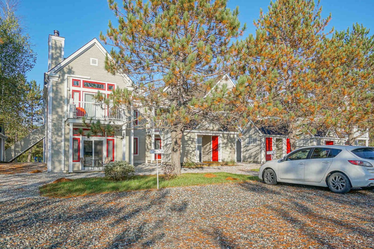 A two-story building is surrounded by trees and a gravel pathway. Bright red doors complement the neutral exterior. A parked silver car is visible in the foreground, while a staircase leads to the second floor. The overall setting is peaceful and well-maintained.