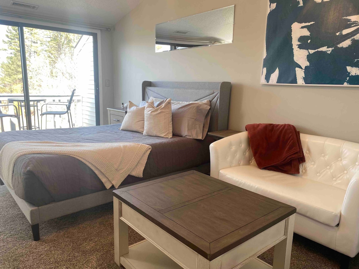 A spacious studio unit features a comfortable bed with soft linens and a neutral duvet. A white upholstered couch is positioned beside a dark wood coffee table. Natural light enters through large glass doors, leading to a balcony with outdoor seating.