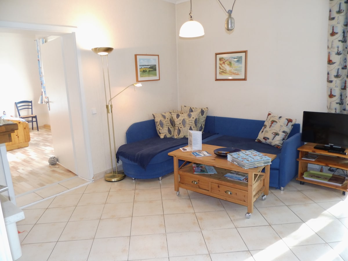 A comfortable living area features a blue sectional sofa adorned with patterned cushions. A wooden coffee table with wheels is positioned in front, while a television is set on a wooden stand. Light is provided by a tall floor lamp, and nautical art decorates the walls.