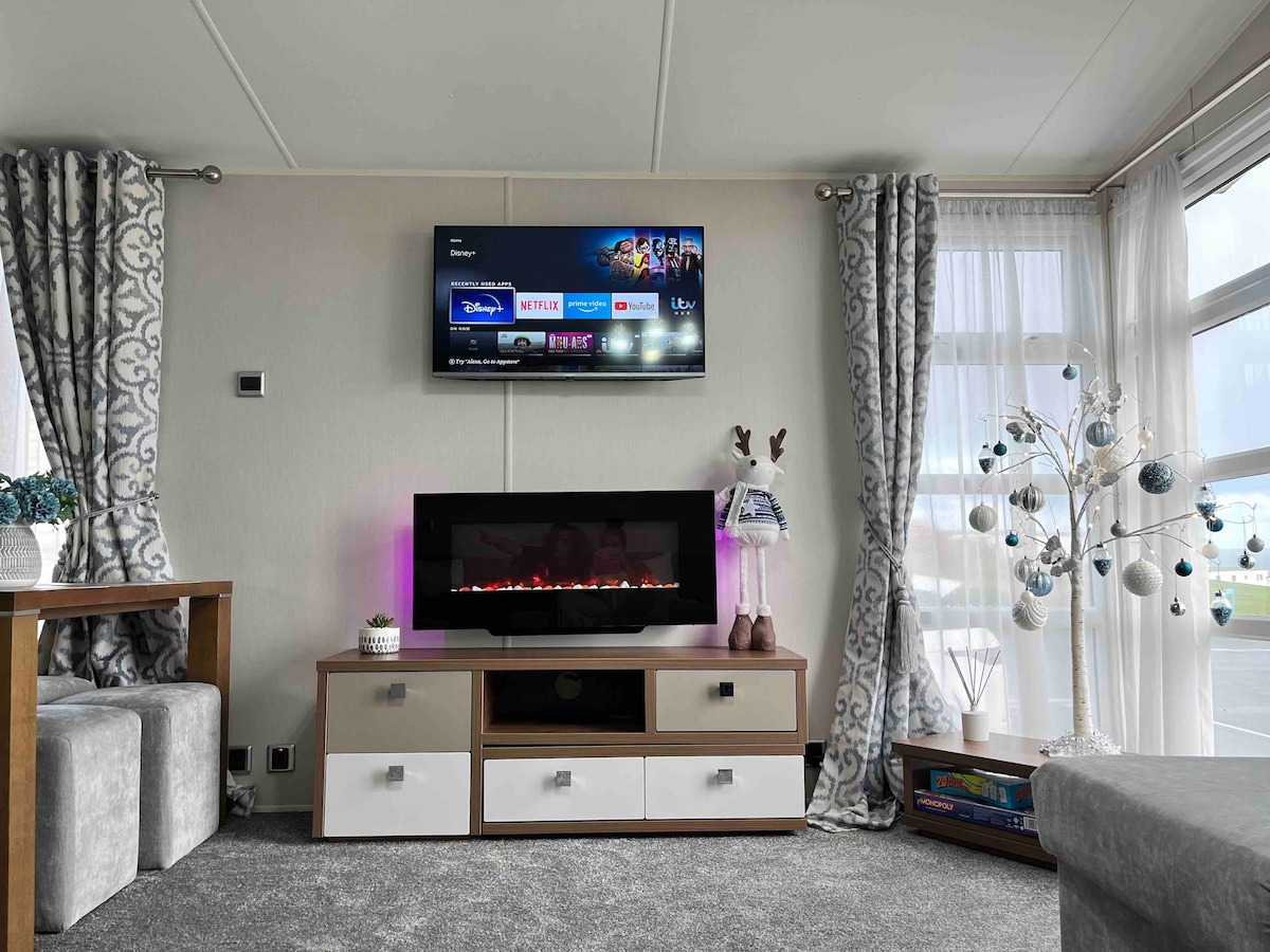 The living area features a wall-mounted TV above a modern media console. A fireplace adds warmth to the space, and decorative curtains frame the large windows, allowing natural light to fill the room. A small, styled tree with ornaments enhances the welcoming ambiance.