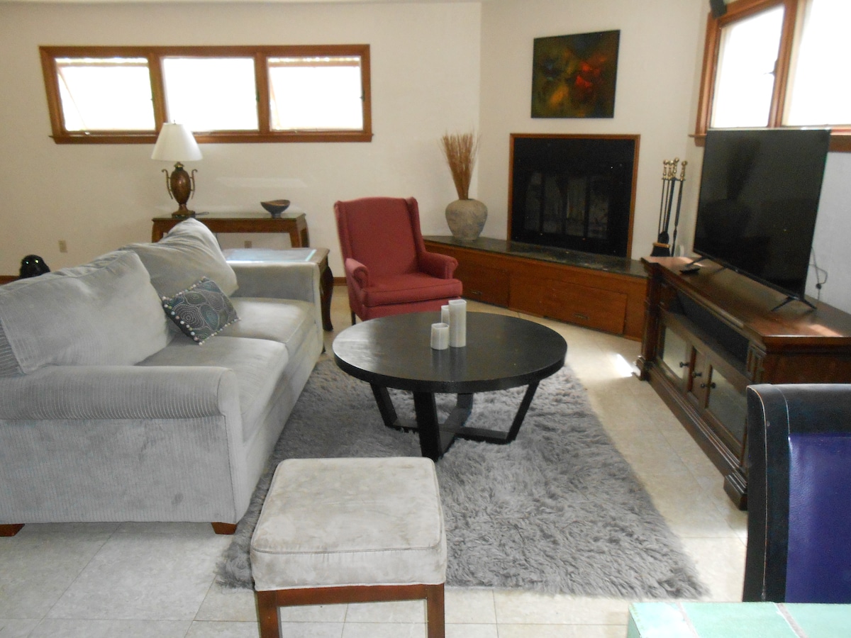 The spacious living room features a comfortable grey sofa alongside a red accent chair. A round black coffee table is centered on a soft area rug. A television is positioned on a wooden console, with natural light filtering in through multiple windows.