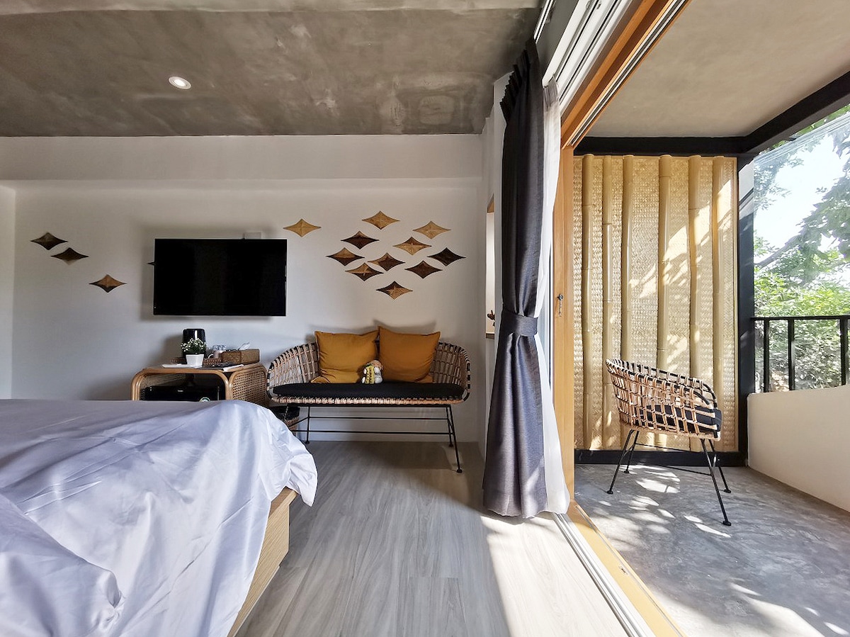 A modern indoor space features a spacious bed with crisp white linens. A television is mounted on the wall, while a cozy seating area with decorative cushions is visible. Natural light streams in through large glass doors, leading to a balcony with views of greenery.