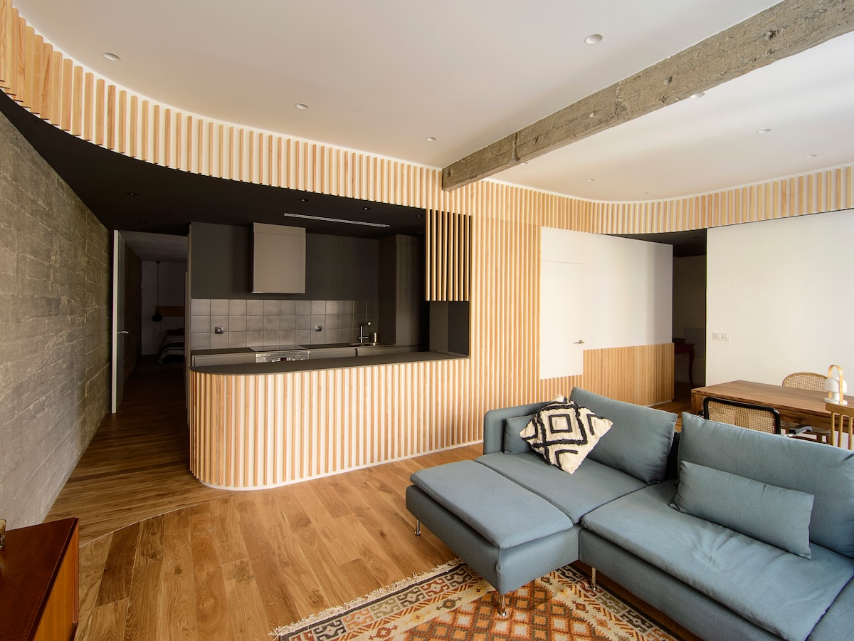 An open living space features a modern kitchen with a sleek countertop and wood paneling. A comfortable sectional sofa forms the focal point of the room, complemented by a patterned area rug. Natural light illuminates the space, enhancing the warm wooden flooring.