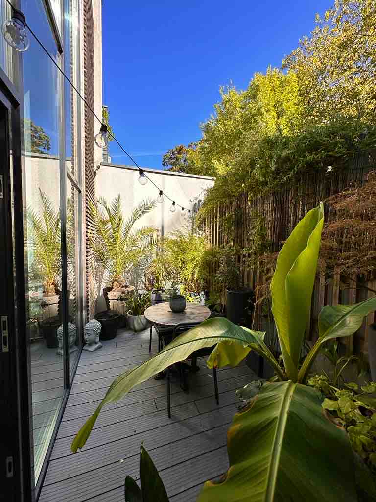A serene patio space is surrounded by lush greenery, featuring a small round table with chairs. Tall plants create a tranquil atmosphere, while string lights offer gentle illumination. The bright blue sky adds to the inviting feel of this outdoor area, promoting relaxation.