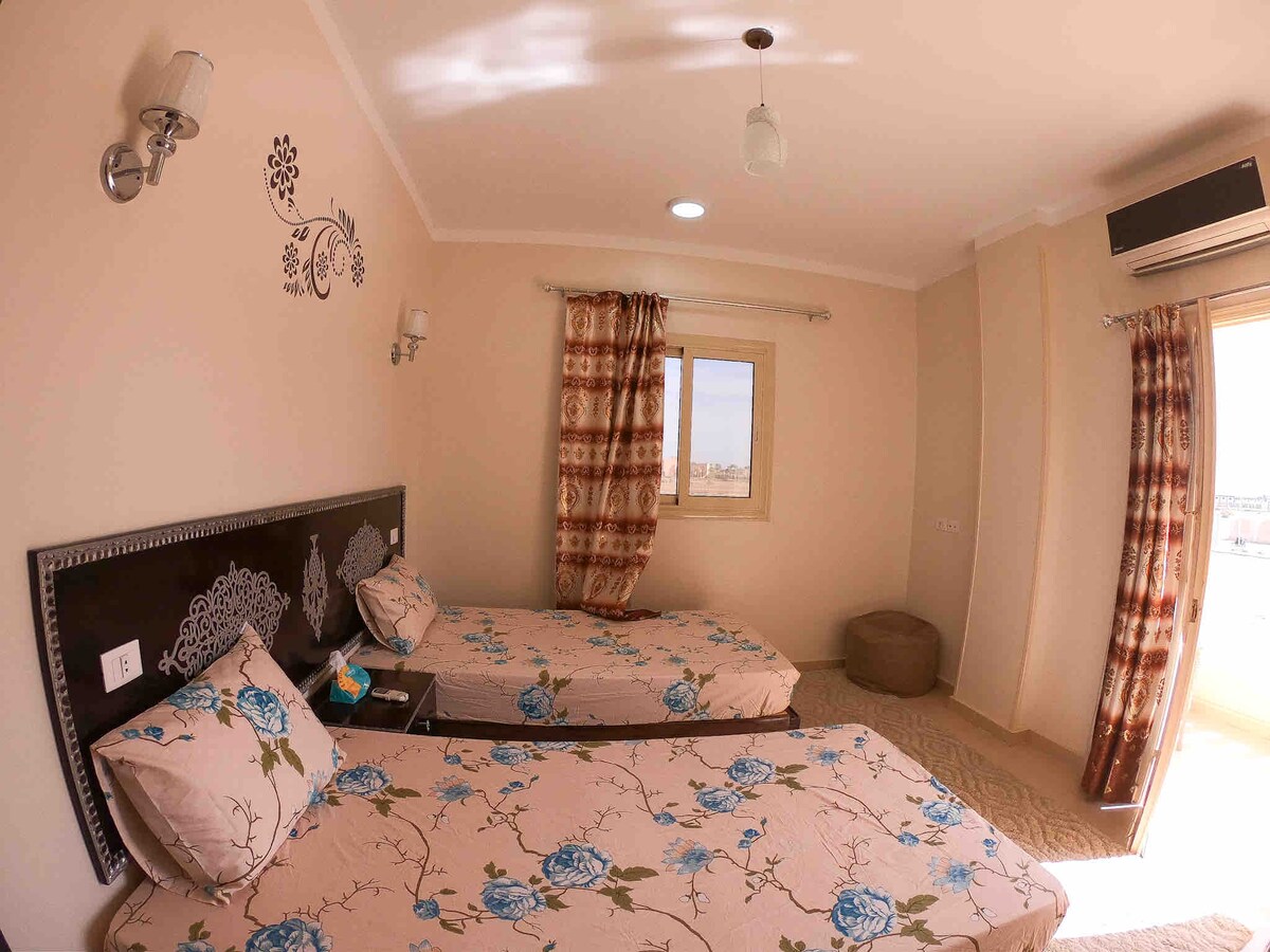 A room is furnished with two cozy beds featuring floral-patterned linens. Soft, neutral walls are complemented by decorative wall art. Natural light enters through a window adorned with patterned curtains, while a climate control unit is mounted on the wall.
