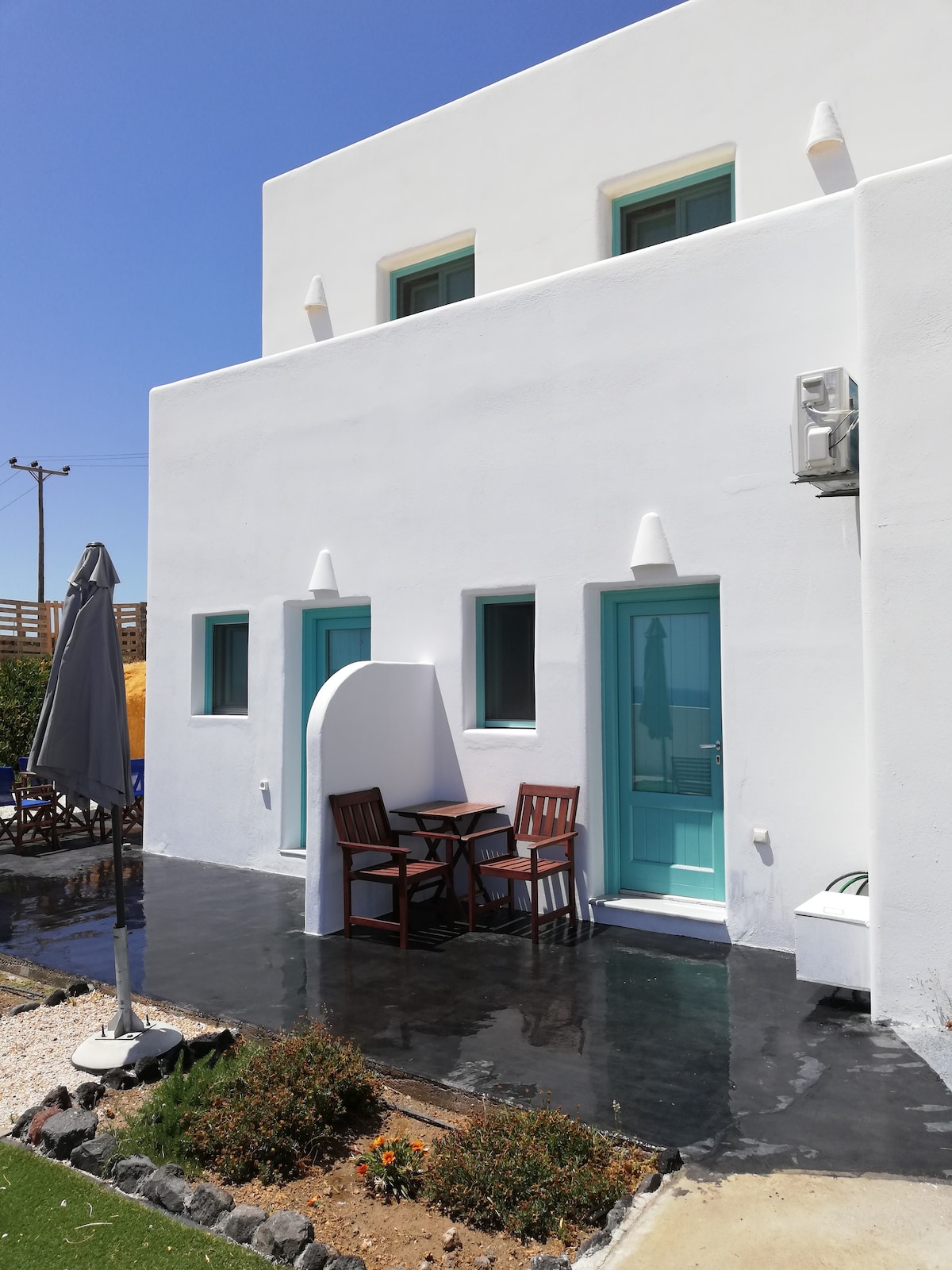 An exterior view of the building showcases white walls accented by teal doors. A compact outdoor seating area features a small table and two wooden chairs. The space is surrounded by a well-maintained garden area, enhancing the inviting setting.