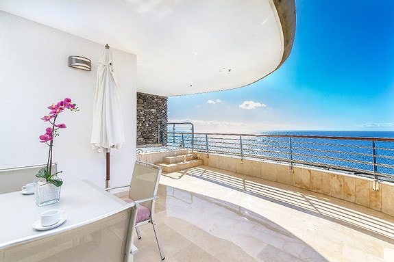 An outdoor terrace overlooking the ocean features a dining table with seating for four. A decorative orchid sits on the table, while a shaded area is provided by a sun umbrella. The glistening water of the sea is visible in the background.